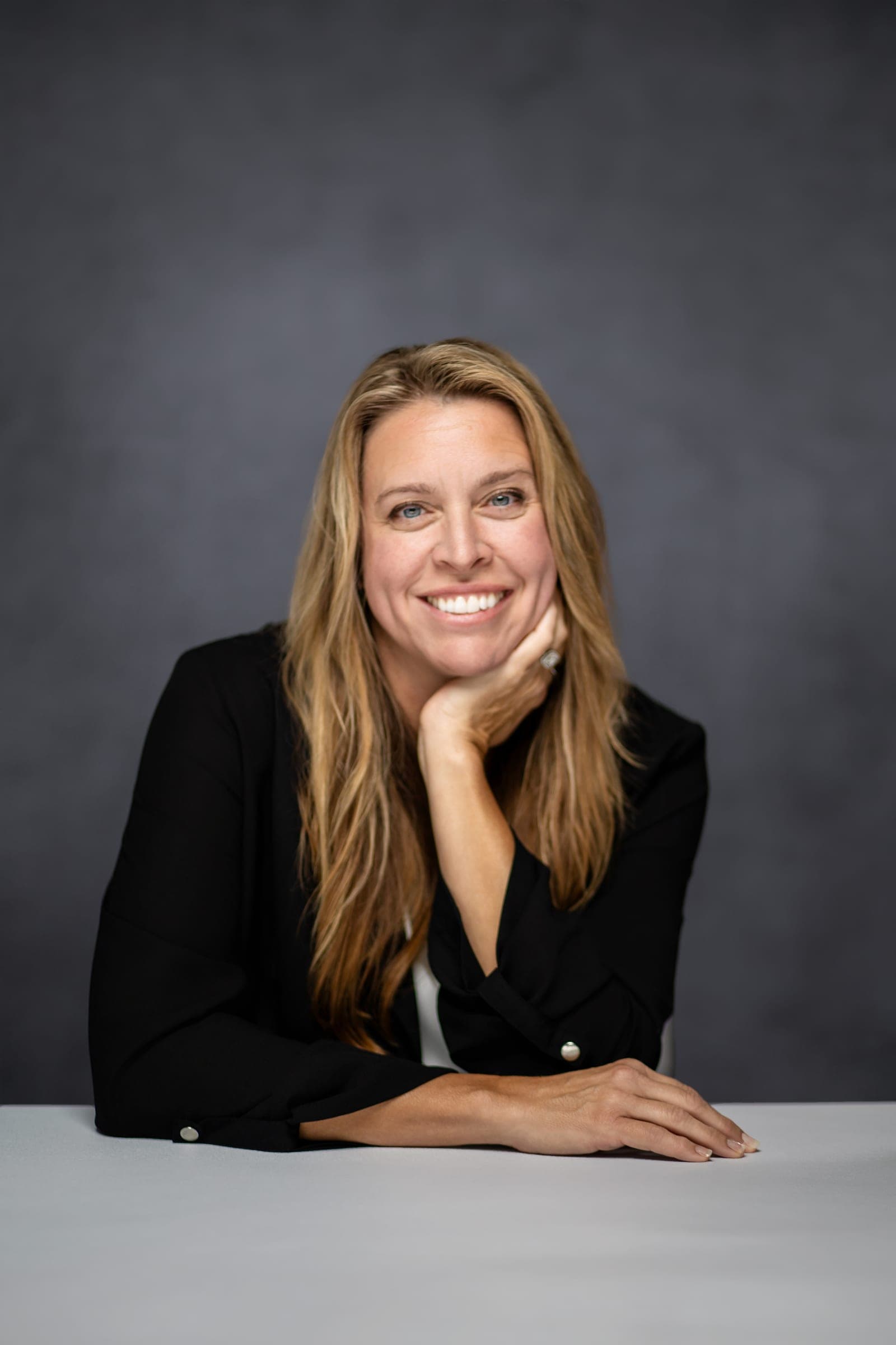 Julie Wilcoxson smiling, chin resting on her hand, in a black blouse at a white table